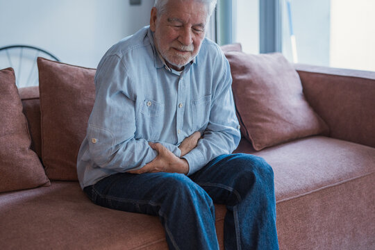 One old man feeling bad and suffering stomachache at home sitting on sofa massaging and touching belly.