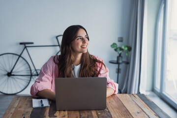 A dreamy young woman sits at her desk with a laptop, gazing into the distance while taking a break from work, reflecting on ideas and visualizing future plans in a calm and comfortable home