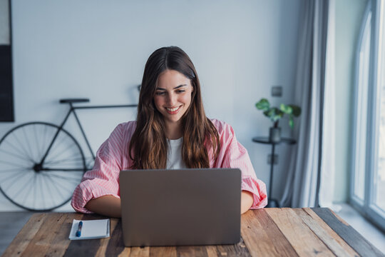 Happy young woman sit at desk look at laptop screen browsing surfing wireless Internet at home, smiling millennial female employee work on modern computer in living room, technology concept