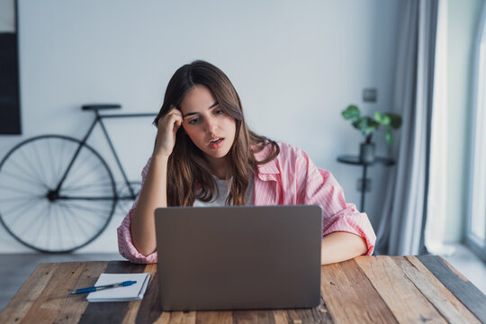 An overworked woman sits at her workspace beside a laptop, pressing her hands to her head as she reacts to negative news from work or bank emails, showing anxiety and emotional exhaustion