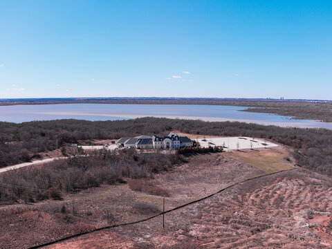 Aerial view of a grand building nestled amidst a forest near a tranquil lake, with a distant cityscape shimmering on the horizon, Denton, Texas, United States.