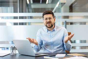 Businessman looking confused and shrugging at a laptop, experiencing a technical issue or...