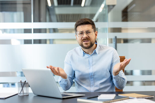 Businessman looking confused and shrugging at a laptop, experiencing a technical issue or misunderstanding while working in a modern office with glass walls