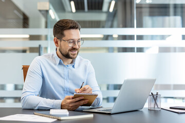 Obraz premium Young businessman wearing glasses sitting at a desk, looking at his laptop screen and writing diligent notes in a spiral notebook, showcasing productivity in a modern office environment
