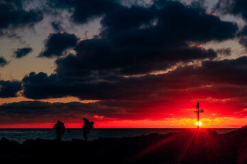 Two silhouetted figures walk along a rocky gates to Klaipeda port under a dramatic red sunset sky, with a distant navigation beacon on the horizon creating a moody, cinematic coastal scene. 