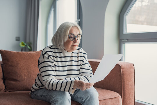 Focused elderly 60s woman sit on sofa in living room reading postal correspondence or bank notice, concentrated mature 50s female rest on couch at home consider message in paper post letter
