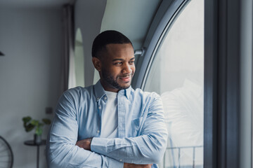 Fototapeta premium Smiling dreamy African American man thinking about future or new opportunities, positive pensive businessman at window looking to aside, planning, pondering strategy, business vision concept