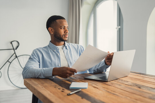 A concentrated African American manager looks out the window while holding a letter and company reports, thoughtfully analyzing statistics, financial risks, and a potential business offer in office