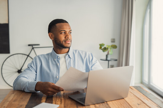 An African American professional gazes out the window while holding business documents, reflecting on company performance, financial risks, and strategic decisions in a thoughtful workplace moment