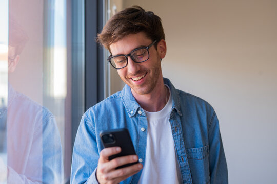 A happy young Latin man stands by the office window typing on his smartphone, sending messages and handling online communication while managing business proposals in a modern workplace setting