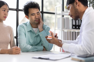 Man with toothache consults with doctor in examination room. Dentist demonstrating an anatomical model of the human jaw while explaining treatment options to a patient in a dental clinic.