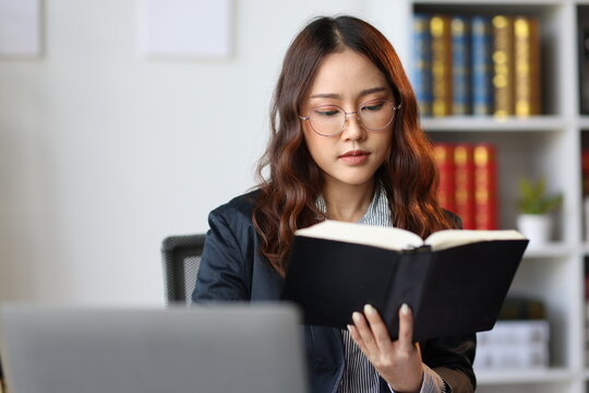 Asian female lawyer reading a law book and studying a case sitting at her desk in a law firm or legal office, she is wearing glasses and turning a page of the book.