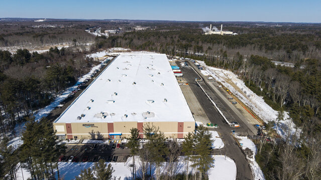 Aerial view of a large rectangular warehouse with snow-covered roof amid a forest, a stark contrast to the clear blue sky, Bellingham, Massachusetts, United States.