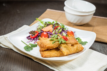 A white plate with golden fried spring rolls served alongside fresh salad greens, cherry tomatoes, and herbs.
