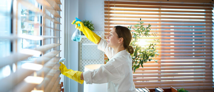 A woman cleans window blinds at home with a rag and gloves, focusing on dust removal, hygiene, household chores, tidiness, daily maintenance, and creating a neat living environment