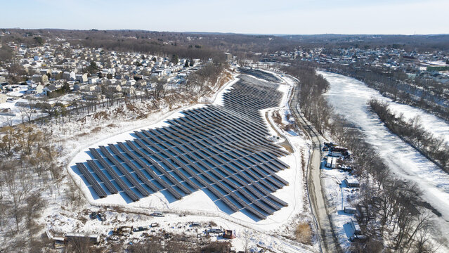 Aerial view of solar panels glinting under the winter sun, nestled beside the winding Blackstone River, a stark contrast against the snow-dusted landscape, Cumberland, Rhode Island, United States.