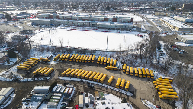 Aerial view of rows of yellow school buses contrast against the stark white snow-covered field and the multi-story building, Providence, Rhode Island, United States.