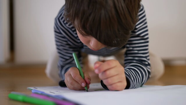 Young boy kneeling on floor, leaning over drawing with green pencil, deeply engaged in creative activity, expressing concentration, focus, and imagination