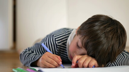 Young boy lying on floor, propped on one arm, drawing with blue pencil, feet playfully raised,...