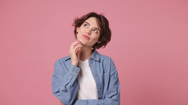 Young dreamful fun woman wears blue shirt white t-shirt casual clothes look around think dream put hand prop up on chin lost in thought and conjectures isolated on plain pastel light pink background
