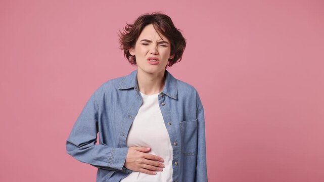 Sick ill sad tired young woman wears blue shirt white t-shirt casual clothes put hands on abdomen suffering from stomach-ache griping bellyache feel bad seedy isolated on plain pastel pink background