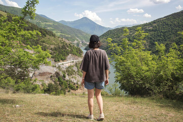Naklejka premium Young woman standing on hilltop overlooking winding river and green mountain valley on sunny summer day, wearing casual clothes and cap, travel and freedom exploration concept