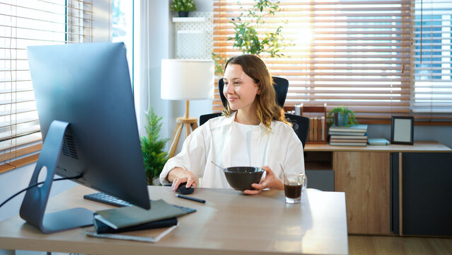 woman sitting at home office table has breakfast with desktop pc. Smiling relaxed young lady hybrid working on computer in modern house