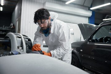 Auto body technician in a protective suit meticulously sands a car hood at a repair shop