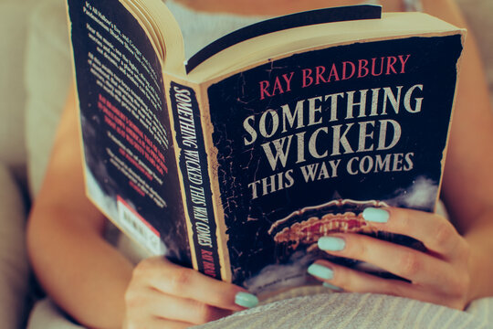 Warsaw, Poland - August 11, 2025: A woman reads a worn paperback copy of Something Wicked This Way Comes by Ray Bradbury. The close-up captures her hands, soft indoor light, and the creased cover.