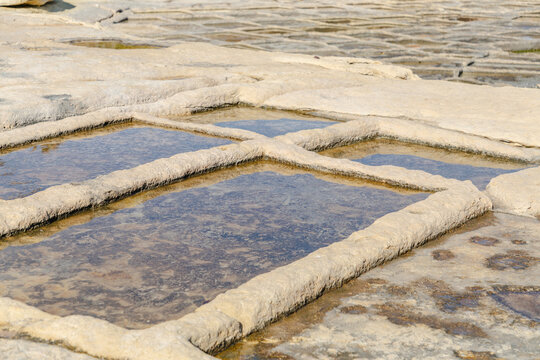View of geometric salt pans filled with shallow water reflecting the sky, carved into the rugged coastline under the sun, Marsaskala, Malta.