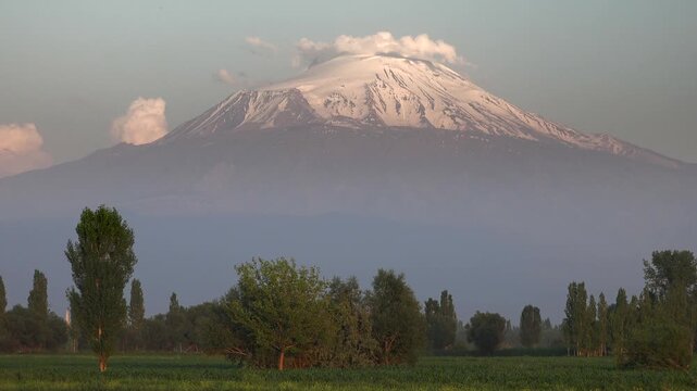 Mount Ararat snowy summit rises above green plain as clouds drift in time lapse sequence. Agri Dagi volcano crown emerges over flatlands, Masis cone glowing under moving sky.