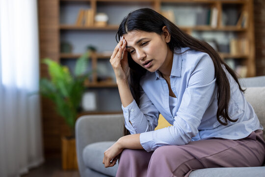 Young woman with dark hair sitting on a sofa at home, holding her forehead and wincing from a migraine or severe headache, tired, stressed and emotionally drained