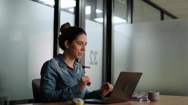 Young business woman in denim shirt smiling while closing laptop at end of workday in contemporary corporate office space