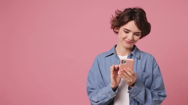 Young amazed woman she wears blue shirt white t-shirt casual clothes hold in hand use mobile cell phone look aside on workspace area copy space mock up isolated on plain pastel light pink background