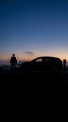 Naklejka premium Silhouette of a man standing by an electric SUV charging at a station against a city skyline at dusk. Represents sustainable energy and future travel. Ideal for automotive concepts.