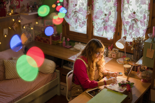 A young woman with curly hair smiles while working on her laptop at a desk in a cozy, decorated room with string lights.