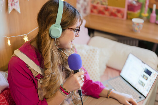 A young woman wearing headphones and glasses holds a microphone while looking at a laptop screen, engaged in a creative activity.