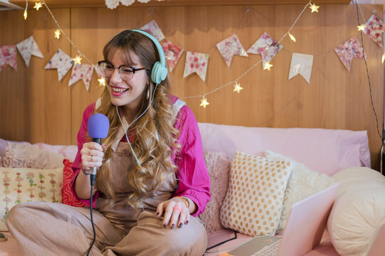A young woman with headphones and glasses smiles while holding a microphone and looking at a laptop, seated on a bed decorated with string lights and banners.