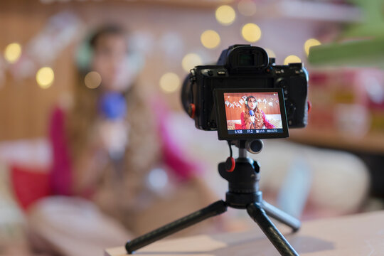 A young woman with curly hair and glasses records a video with a microphone and camera on a tripod, creating content for her online audience.