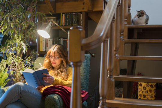 A young woman reads a book in a cozy armchair under a lamp, with a cat perched on the stairs nearby.