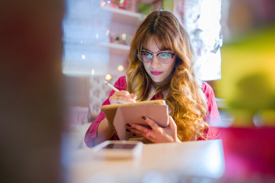 A young woman with glasses is focused on her tablet, using a stylus to draw or write. She is wearing a pink shirt and sitting at a desk.