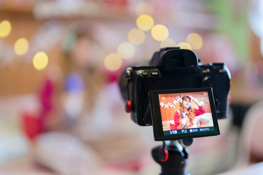 A young woman with headphones and glasses records herself speaking into a microphone, with a camera screen showing her image and festive decorations in the background.