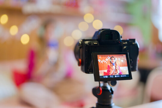 A young woman with headphones and glasses records herself speaking into a microphone, with a camera on a tripod capturing the moment.