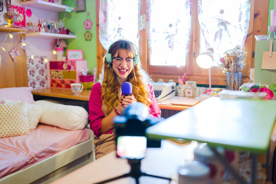 A young woman with headphones and glasses laughs while holding a microphone, recording content in her colorful bedroom.