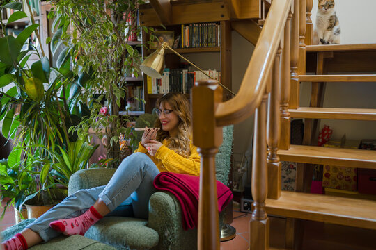 A young woman relaxes in a cozy armchair, holding a phone and smiling. A cat sits on the stairs nearby, observing the scene.