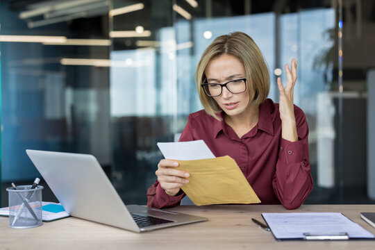Stressed businesswoman reads an official letter at her desk, visibly frustrated and worried in a modern office as upsetting news about work or finances creates anxiety and concern