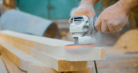 Close-up of hands working to sand wooden beams using an angle grinder. Dust fills the air as the...