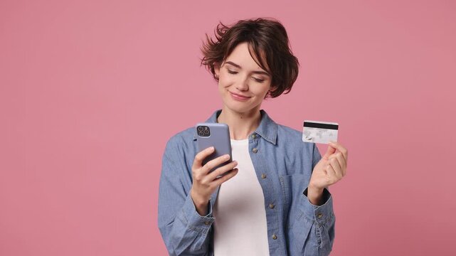 Young woman wears blue shirt white t-shirt casual clothes use mobile cell phone app hold in hand credit bank card doing online shopping order delivery to home isolated on plain pastel pink background