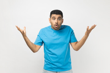 Fototapeta premium Portrait of unshaven man wearing blue T- shirt standing spreads palms, looks astonished at camera, quarrels with someone feels puzzled. Indoor studio shot isolated on gray background.