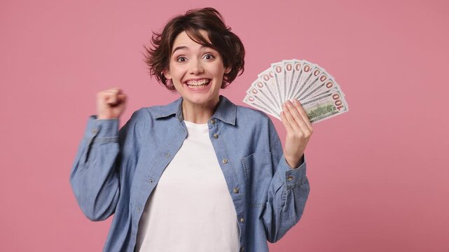 Happy young woman she wearing blue shirt white t-shirt casual clothes hold in hand fan of cash money in dollar banknotes doing winner gesture celebrate isolated on plain pastel light pink background
